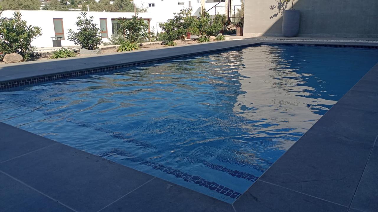 Piscina de hormigón revestida en porcelanato azul profundo con terraza de piedra oscura, construida por Valcorp
